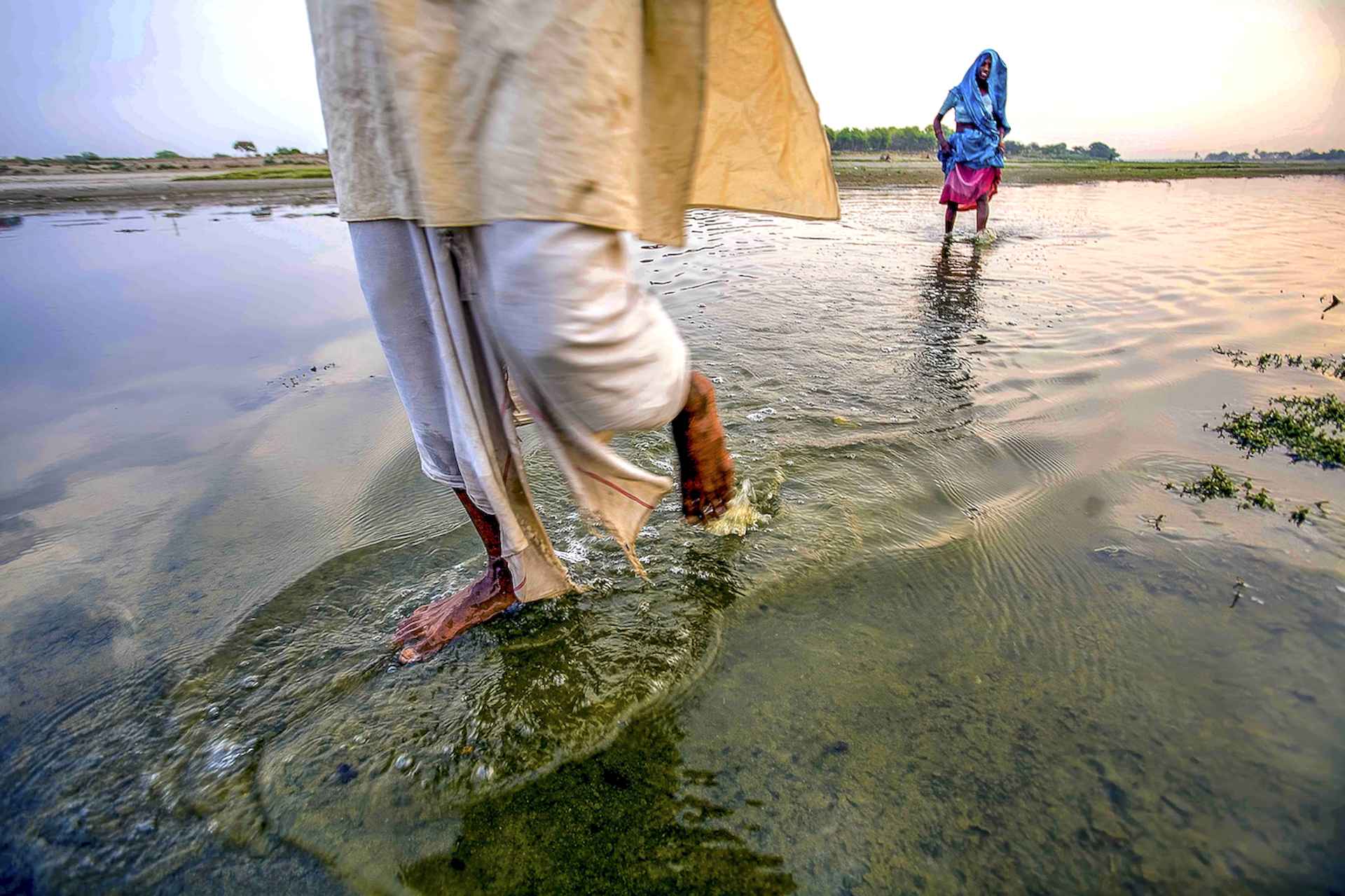 People walking barefoot through shallow river water in traditional Indian clothing at sunrise