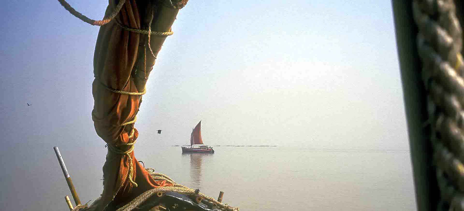View from the bow of a wooden boat on calm water, with a distant sail under soft light.
