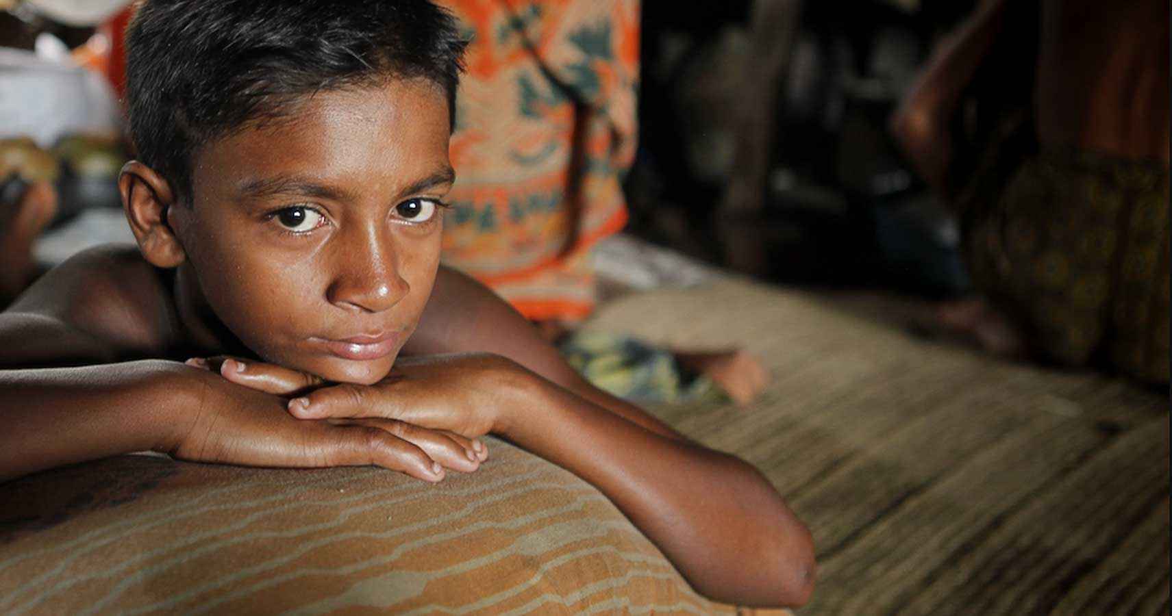 Young boy resting on a cushion inside a modest home, looking directly at the camera, conveying quiet emotion and human connection